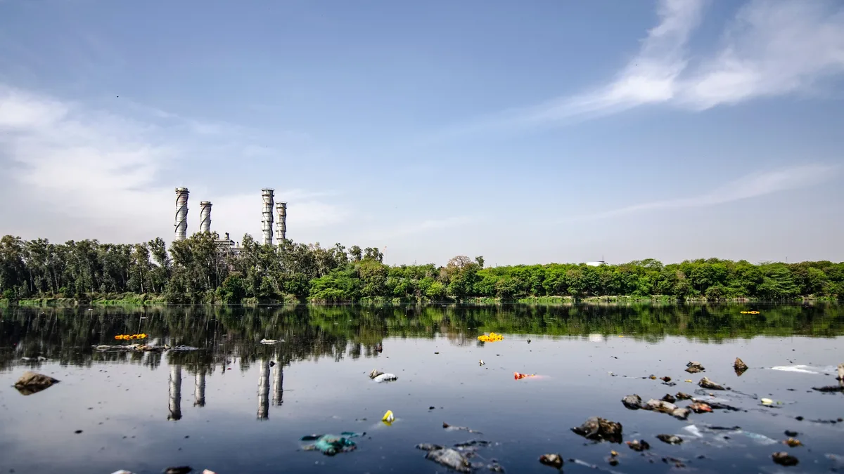 A polluted body of water with scattered trash and debris, set against a backdrop of trees and a blue sky.